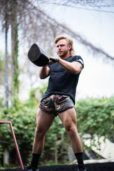 Man lifting a kettlebell outdoors with greenery in the background