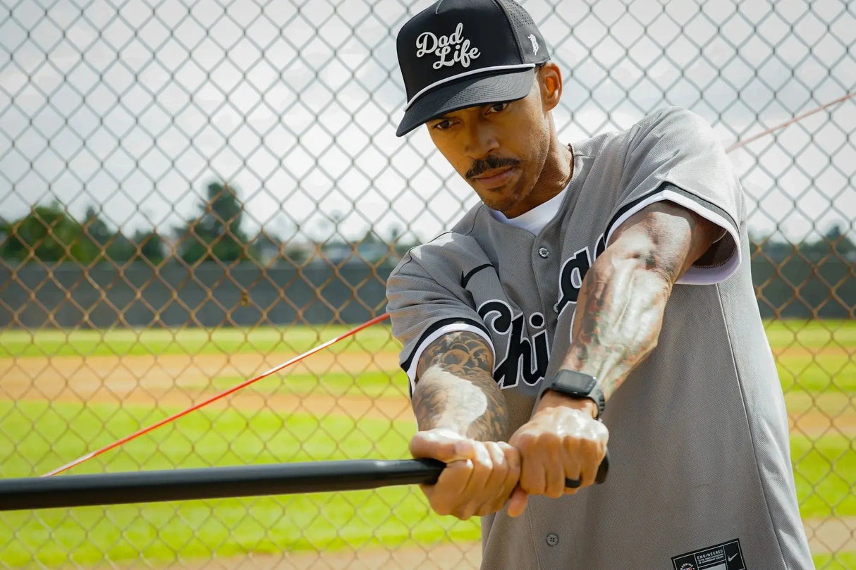 Man in a baseball uniform with 'Dad Life' cap holding a bat on a baseball field.