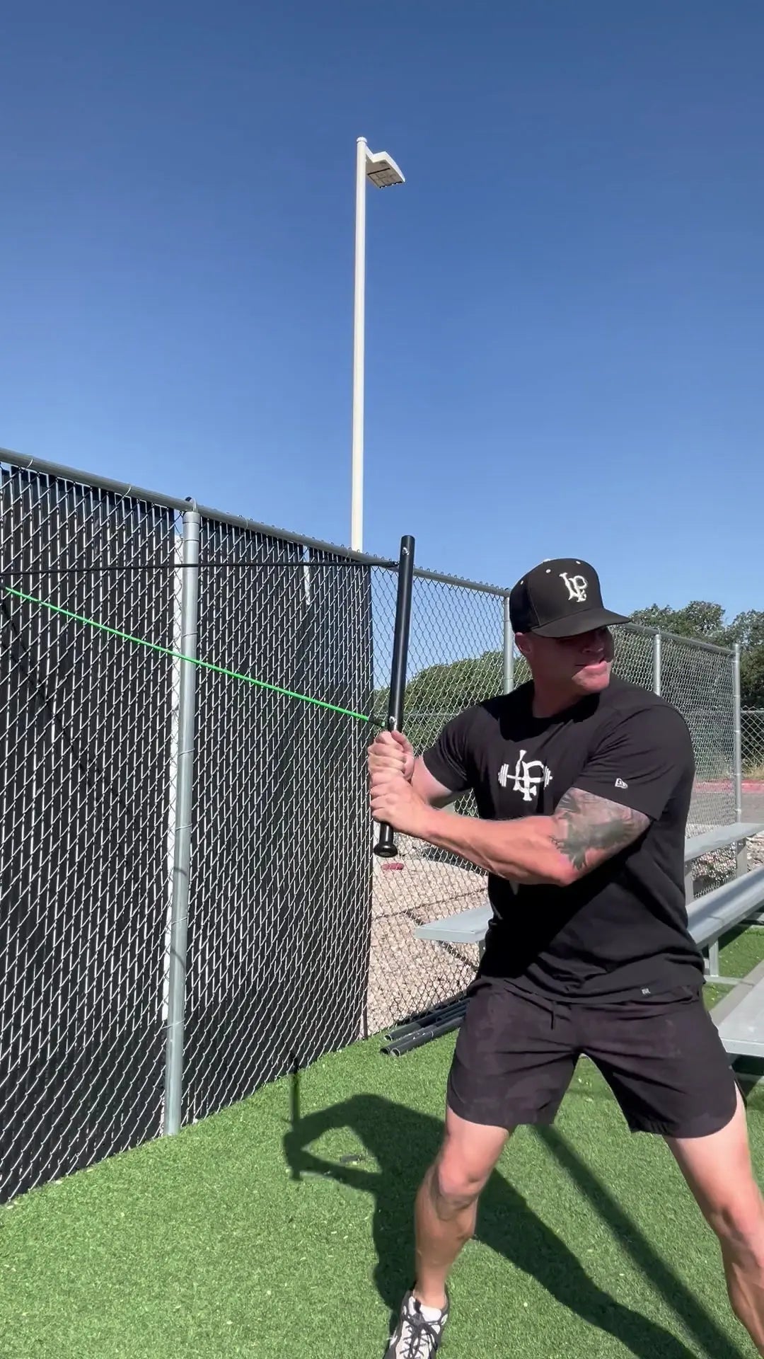 Person in black t-shirt and shorts holding a baseball bat on a sports field with a clear blue sky.