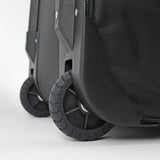 Close-up of a black hard-shell rolling suitcase showing the front wheel, protective wheel cover, and textured fabric side panel on a light gray background.
